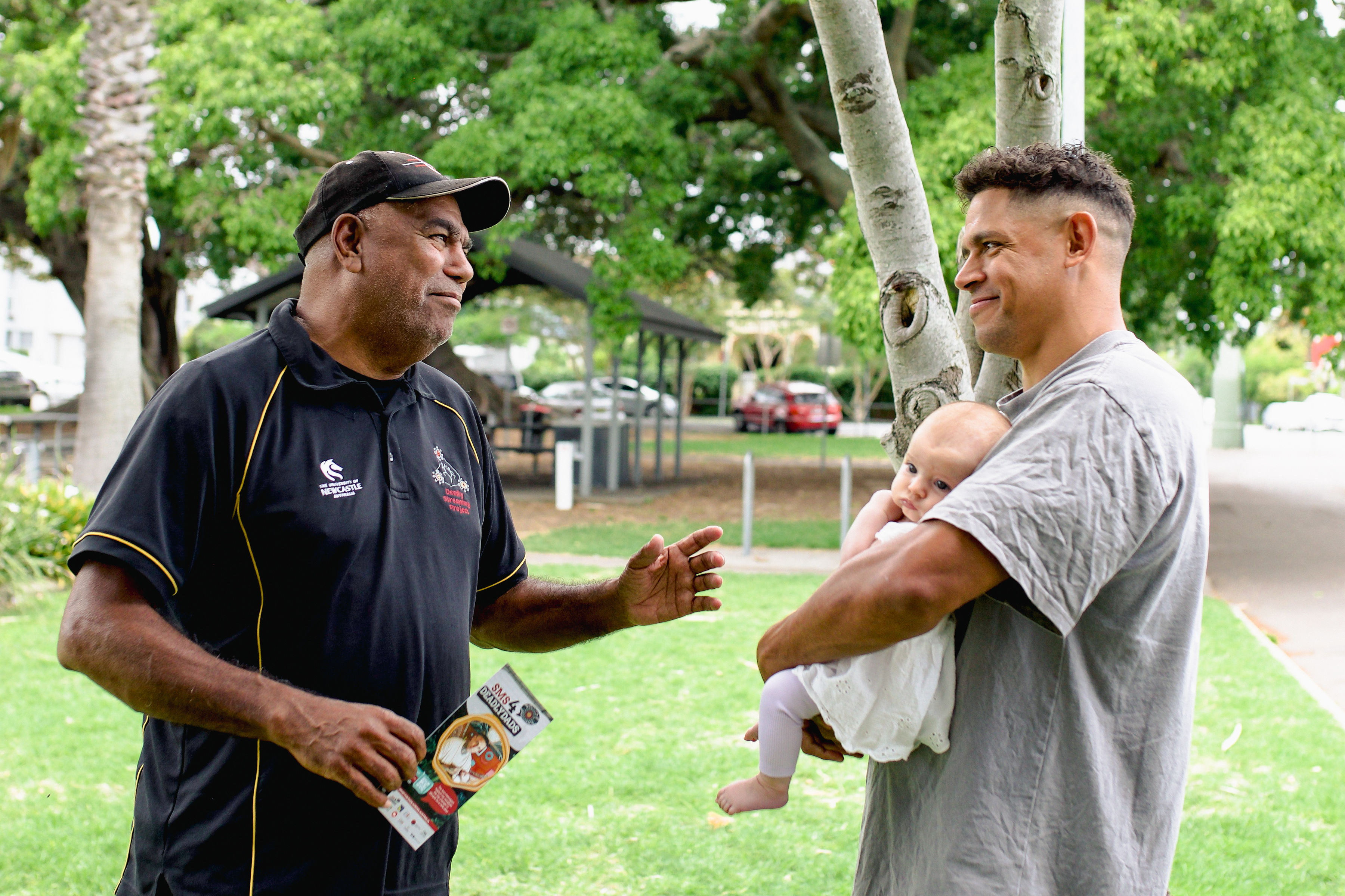 Two men stand smiling at each other. One of the men holds a baby.