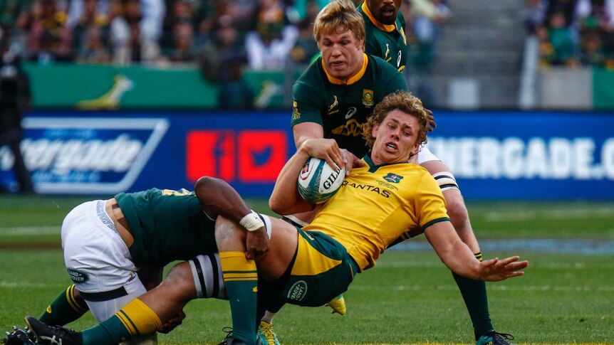 A rugby player in yellow is tackled by two players in dark green