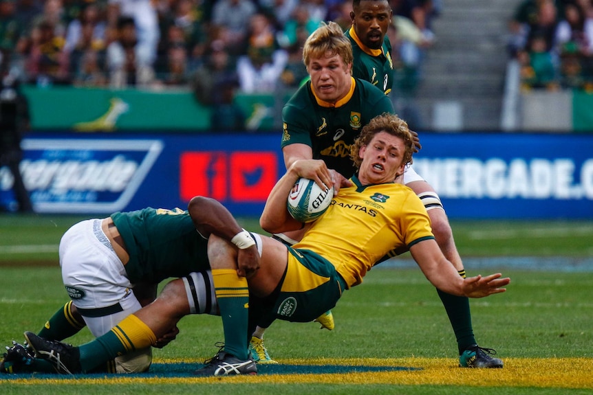 A rugby player in yellow is tackled by two players in dark green