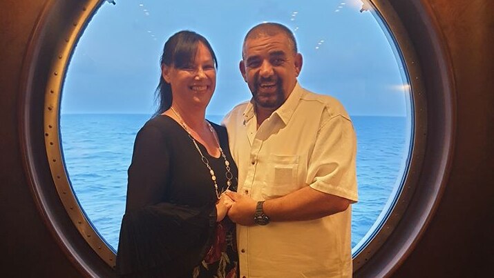 A middle-aged couple smiling and holding each other in front of a huge porthole a cruise ship.
