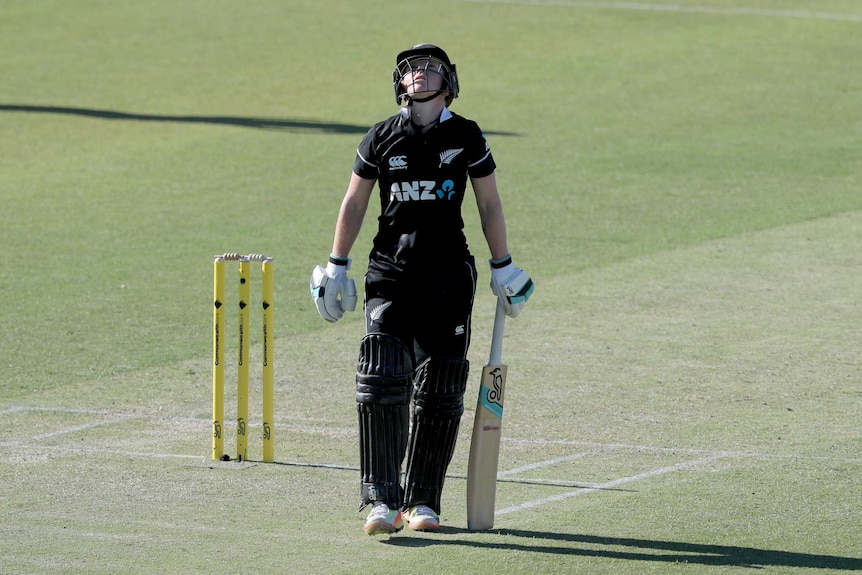 A woman wearing a black NZ cricket uniform, helmet and gloves looks up to the sky on a cricket pitch