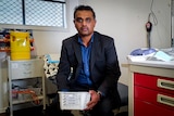 Dr Ranatunga sits in his medical practice, holding a plastic box with COVID vaccines inside
