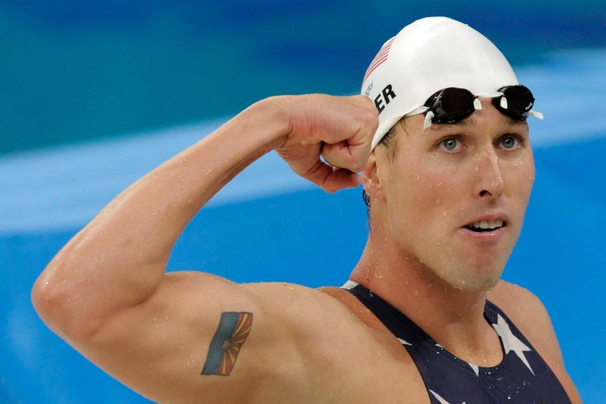 Wet man in white swimming cap, googles and bathers raises right arm in a fist.