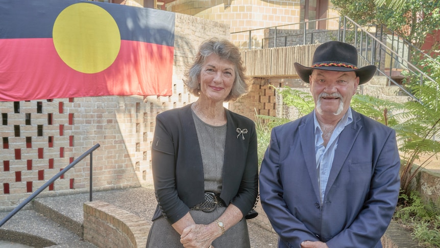 Geraldine Doogue and Jack Beetson pose for a photo in an outdoor courtyard next to an Aboriginal flag