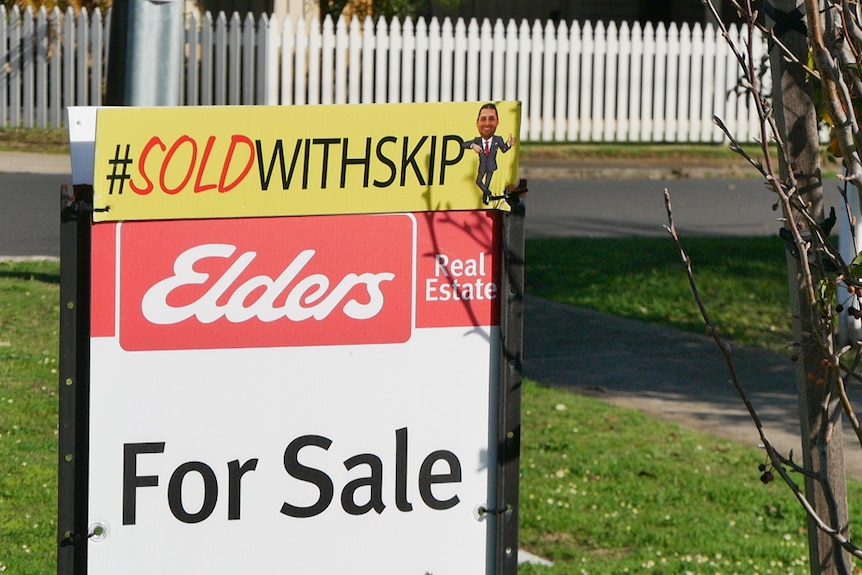 An Elders "For Sale" sign with a 'sold' sticker on the top on the verge of a house.