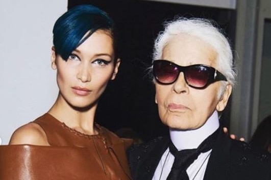 Model Bella Hadid wearing a brown leather dress stands next to Karl Lagerfeld wearing a black suit and sunglasses.