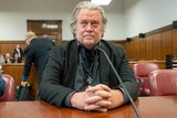 A photo of Steve Bannon sitting at a table in court. He has shoulder length greying hair. 