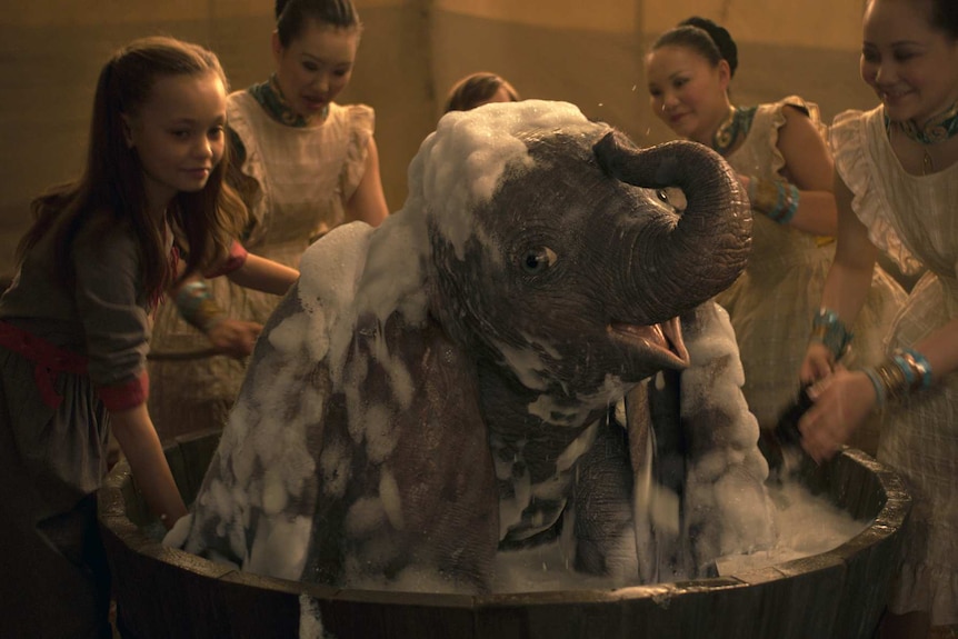 A CGI animated elephant, wreathed in bubble bath bubbles, smiles as a group of women in period garb help him bathe.