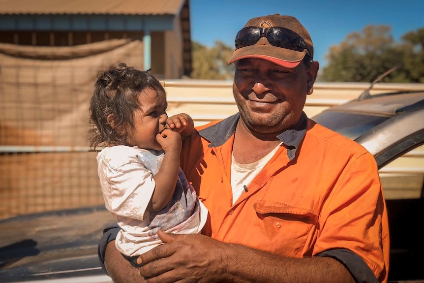 A man in an orange shirt wearing a cap holds a young girl.