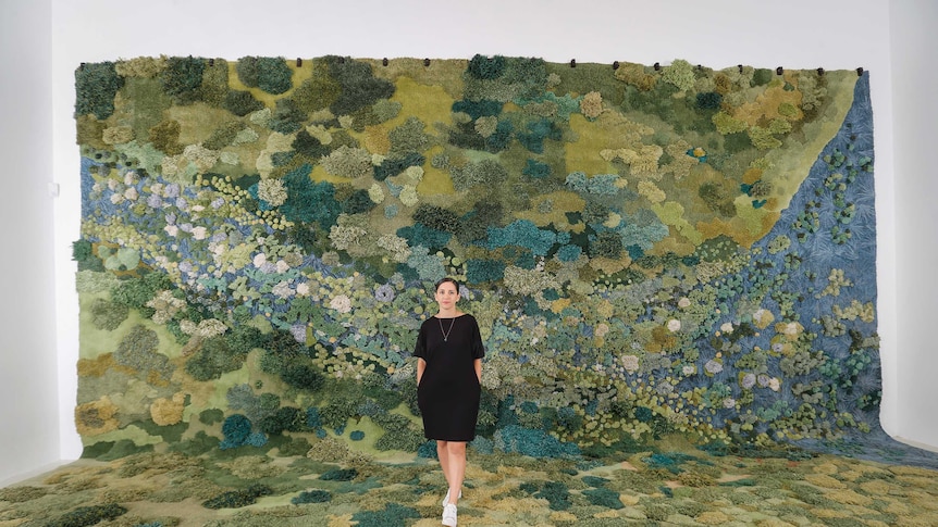 An artist stands in front of a monumental landscape carpet