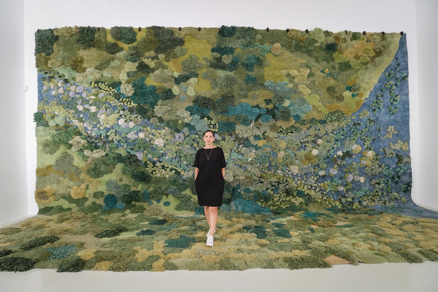 An artist stands in front of a monumental landscape carpet