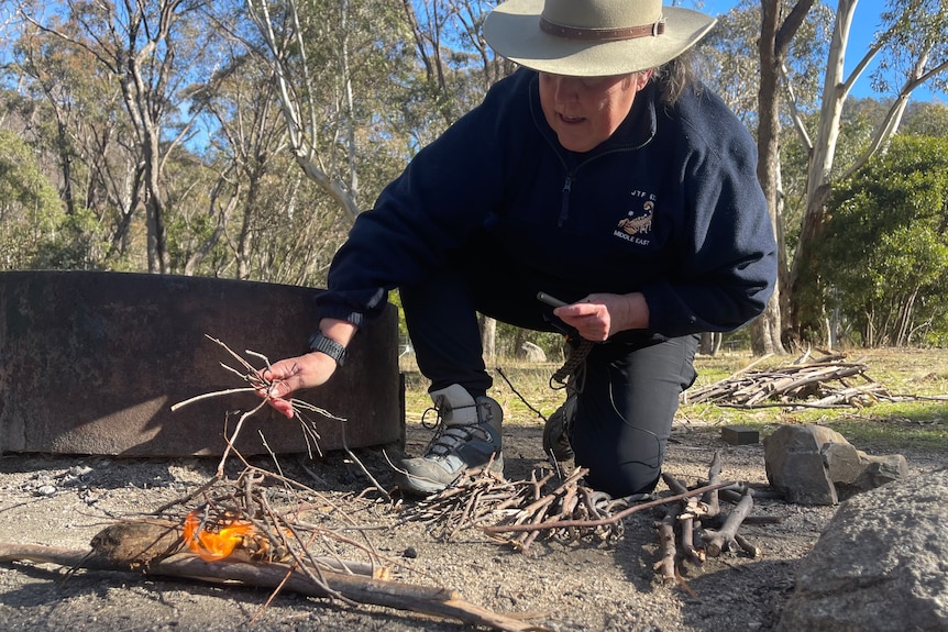 A woman in a broad tan hat bends down while making a fire.