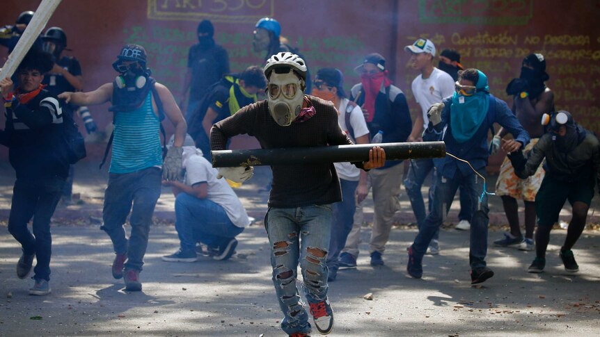 A man wearing a gas mask and bike helmet runs from tear gas holding a homemade mortar.