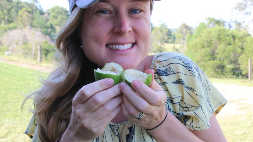 A woman in a cap hold up a split feijoa, smiling