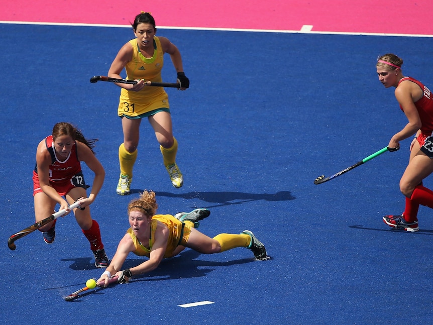 An Australian hockey player sweeps her stick around the ball as she dives full-length on the ground surrounded by US players.
