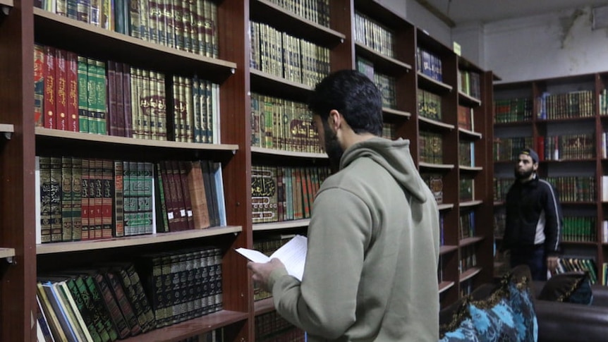 A man stands holding a book facing two walls of tall book shelves, filled with neatly stacked books. Near him stands man.