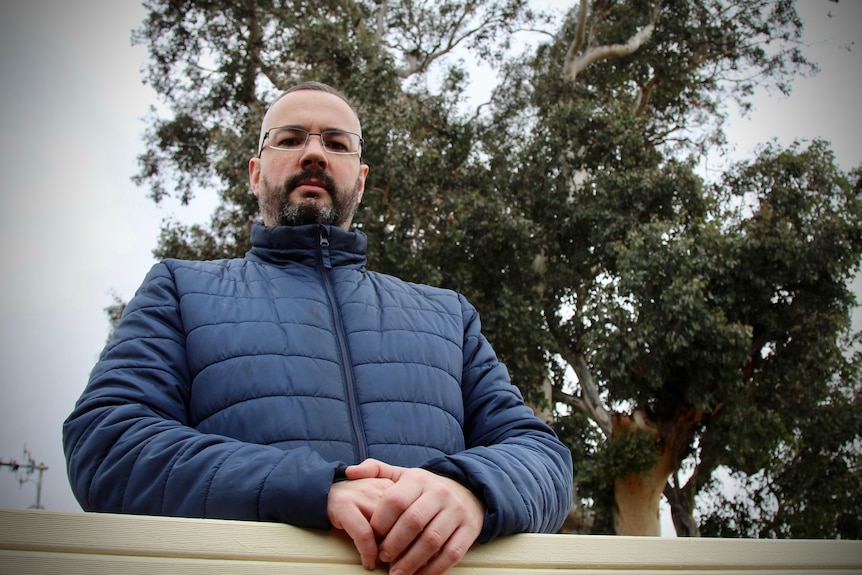 A man in a blue puffer jacket stands in front of a large gumtree and a brick house.