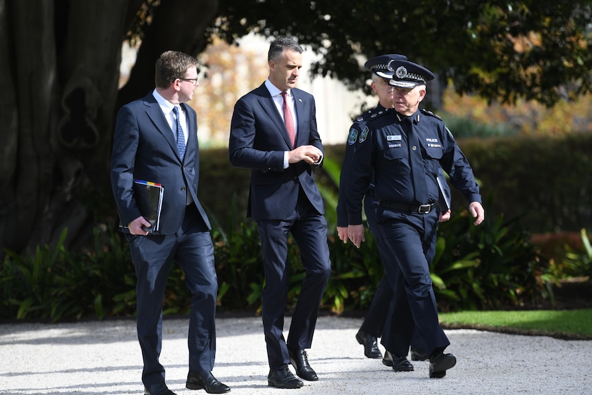 Three men walk along an outside path with a tree in the background