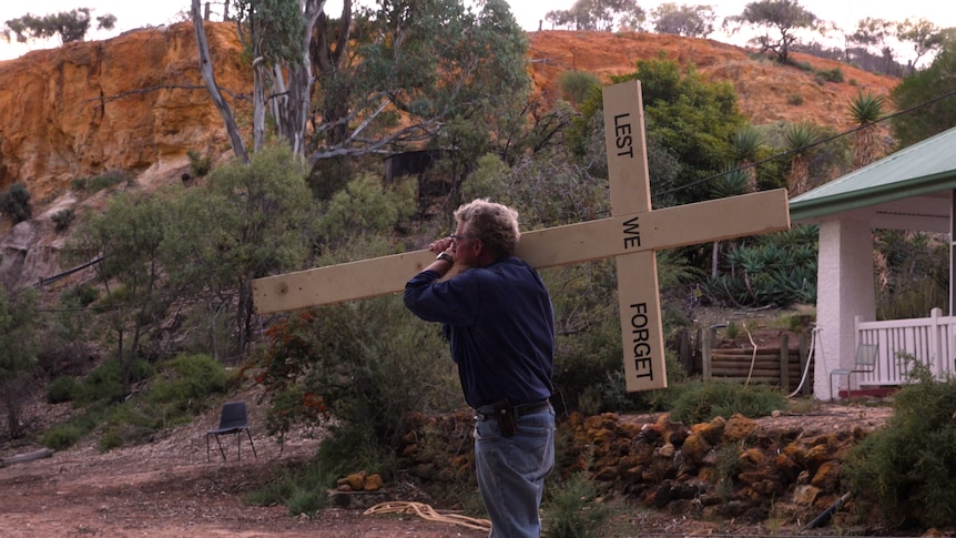 A man with a long beard and white curly hair carries a cross the reads "lest we forget".