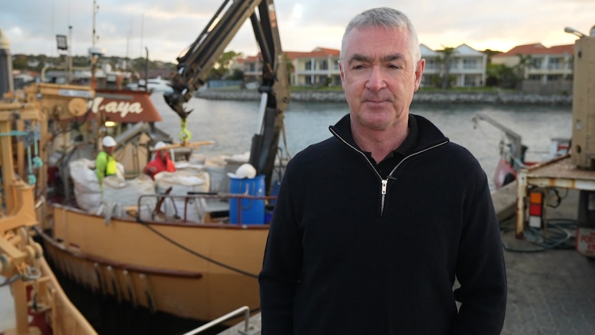 A man with a serious expression stands in front of a boat on the water