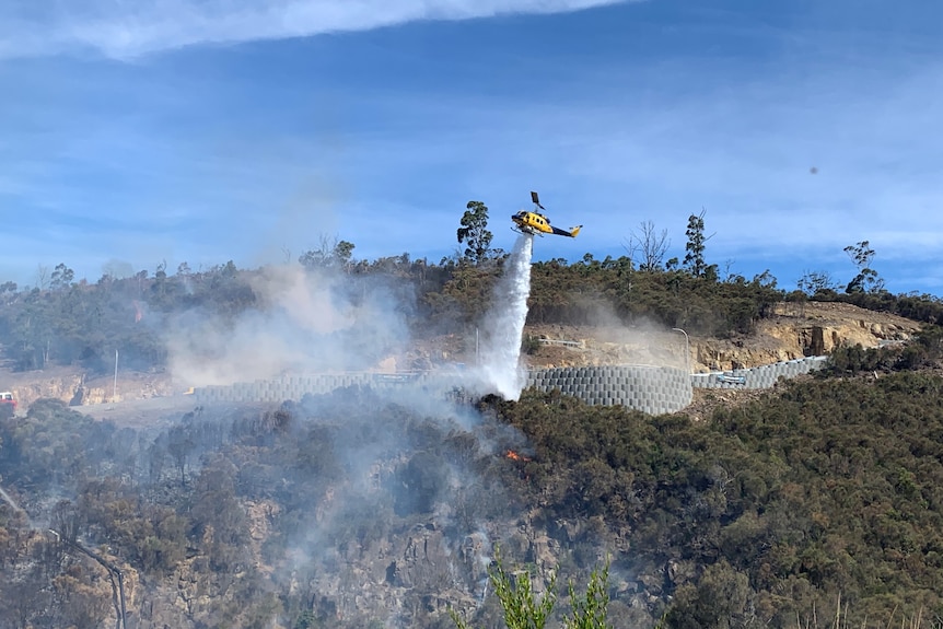 a yellow and blue helicopter can be seen dropping a substance on a blaze with smoke rising from burnt trees on a hill face