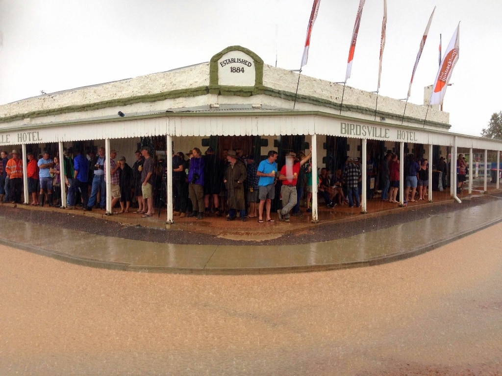A country hotel with patrons standing under the awning outside, holding beers, while it rains.