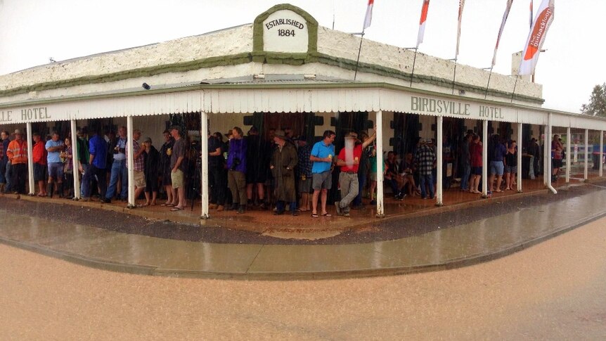 A country hotel with patrons standing under the awning outside, holding beers, while it rains.