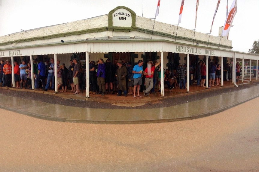 A country hotel with patrons standing under the awning outside, holding beers, while it rains.