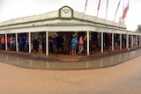 A country hotel with patrons standing under the awning outside, holding beers, while it rains.