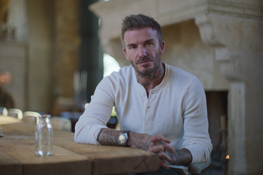 David Beckham, wearing a white long-sleeve t-shirt, sits at a kitchen table looking serious