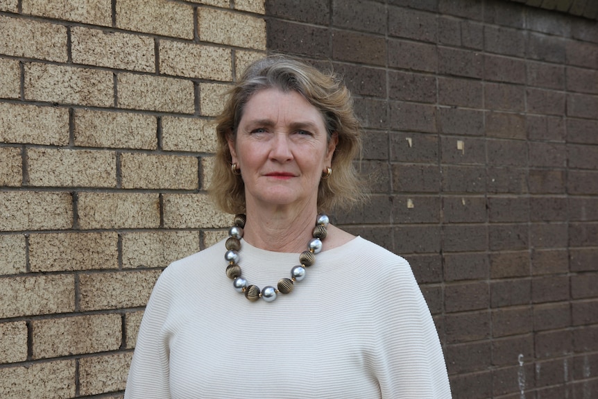 A fair-haired woman wearing a light-coloured top stands in front of a brick wall.