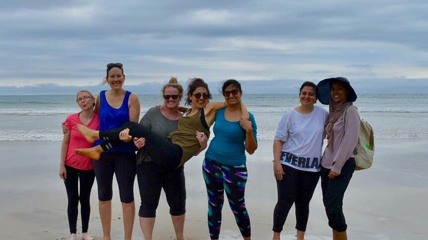 Seven women in exercise clothes stand barefoot on a beach.