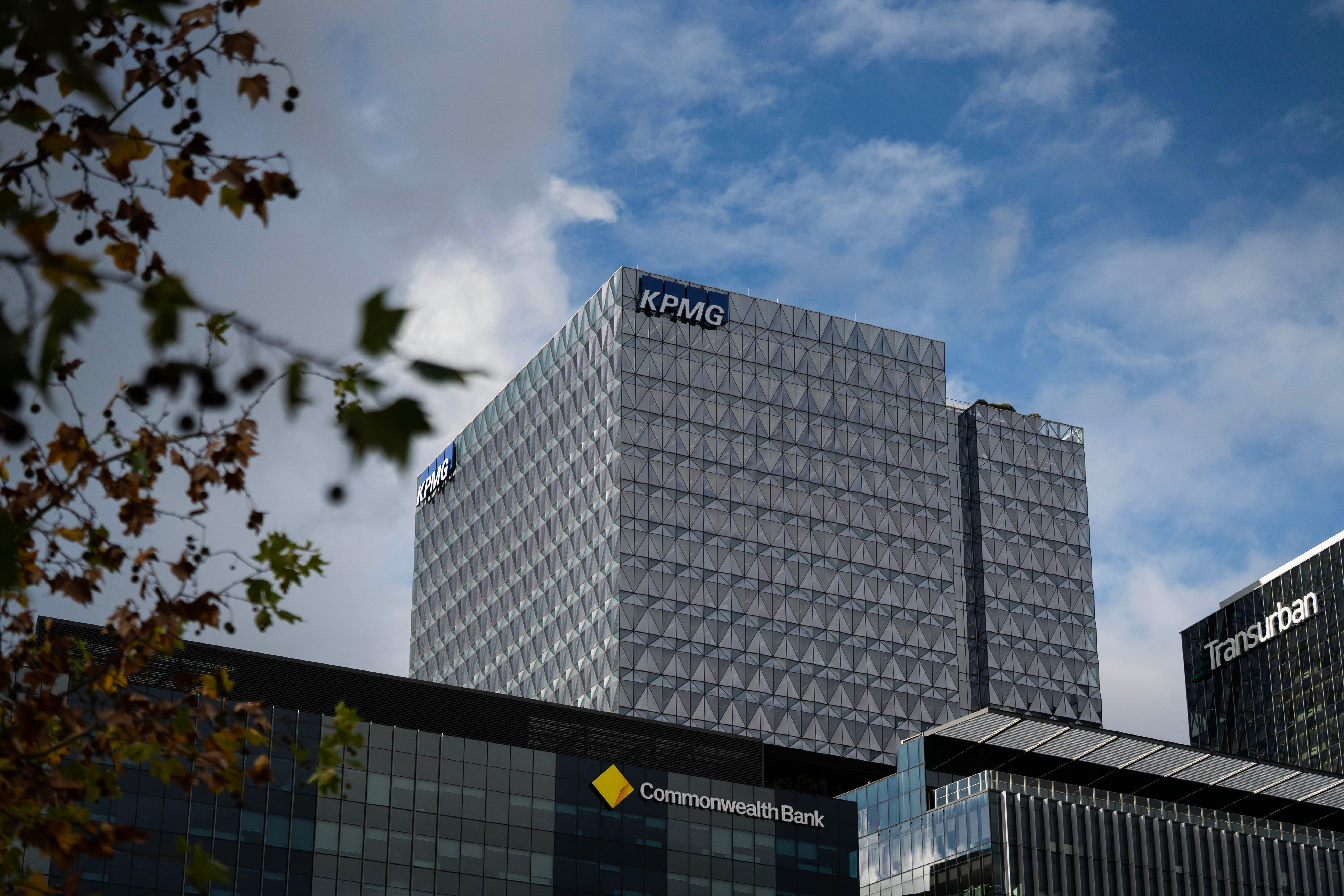 A large, grey building with a blue and white sign at the top reading KPMG. A lower Commonwealth Bank building is in front of it.