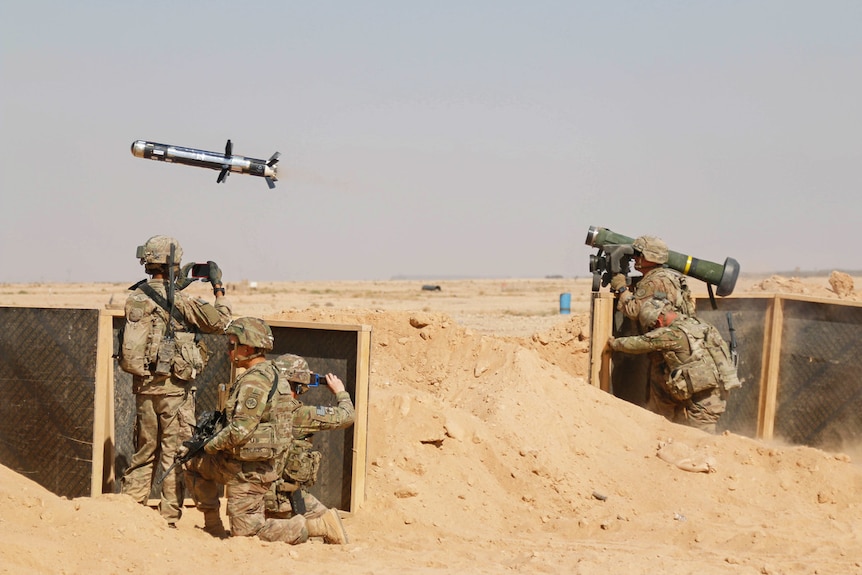 In a desert field, four soldiers look through smartphones and military cameras as a missile fires above their heads.