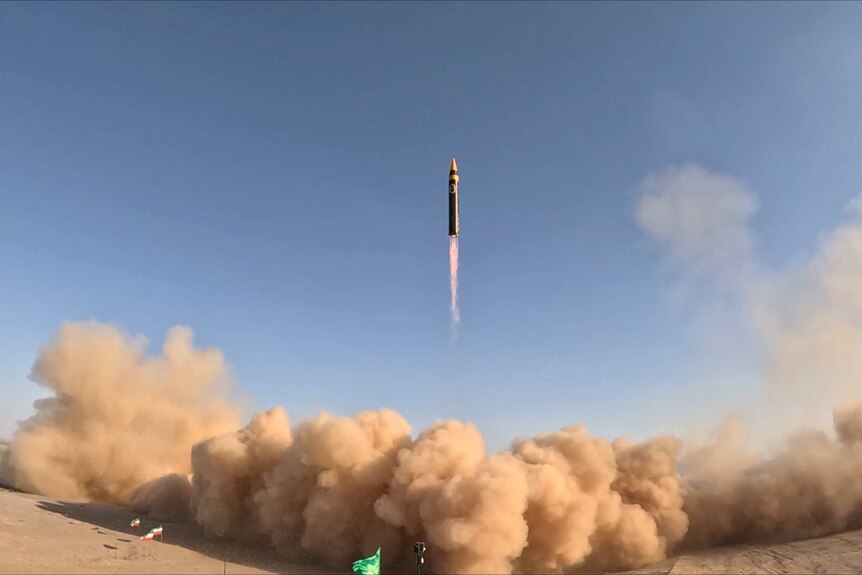 A large black cylindrical ballistic missle launches into the sky with a firey exhaust blowing large clouds of dust.