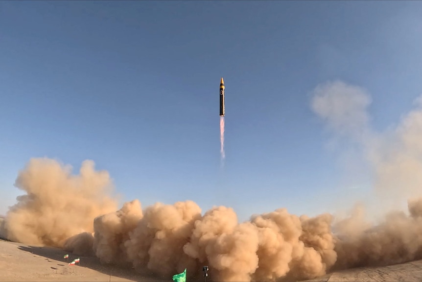 A large black cylindrical ballistic missle launches into the sky with a firey exhaust blowing large clouds of dust.