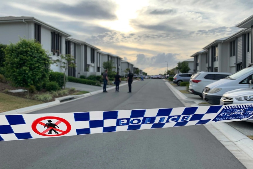 Three police officers stand on a street outside townhouses with crime scene tape in the foreground