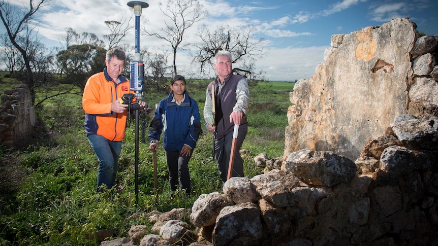 Two men - one on the left with surveying equipment -  and woman in the middle - in a field  next to a big rocky outcrop