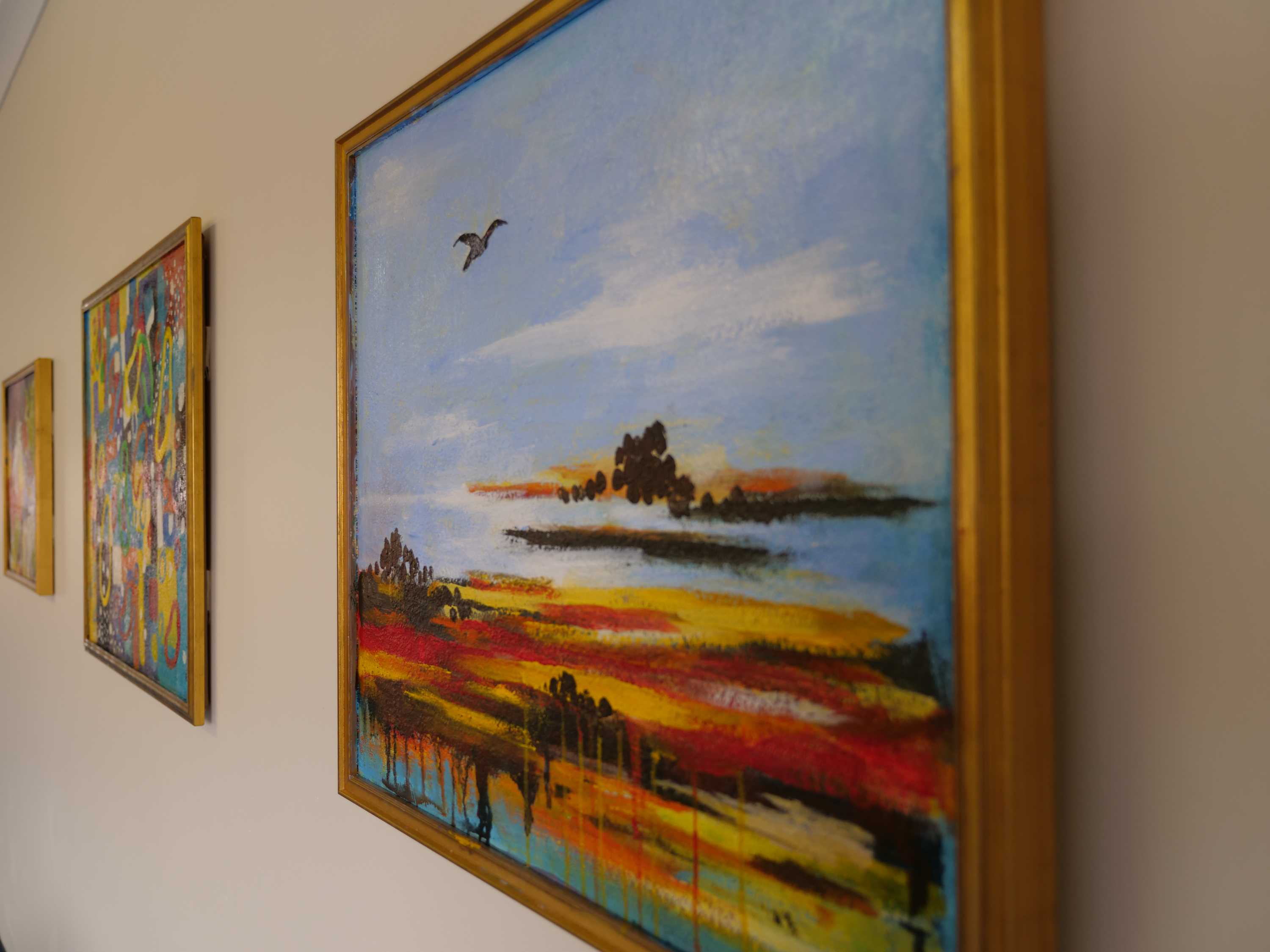 A close up of a landscape painting in a golden frame hanging on the wall, two more paintings are hanging to the left of it.