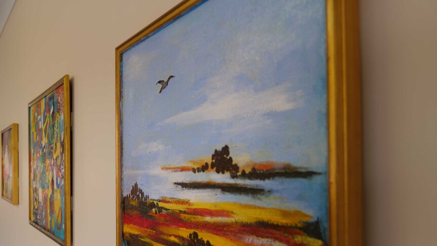 A close up of a landscape painting in a golden frame hanging on the wall, two more paintings are hanging to the left of it.