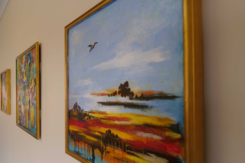 A close up of a landscape painting in a golden frame hanging on the wall, two more paintings are hanging to the left of it.