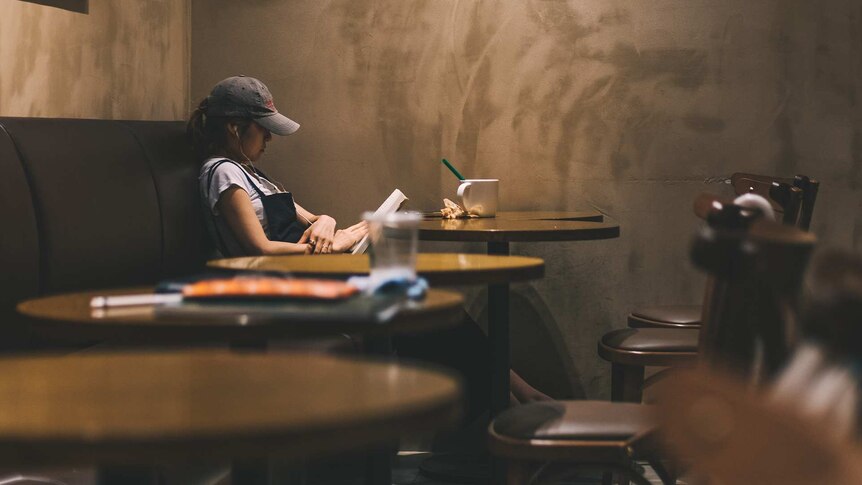 A young woman wearing a cap sits at a dark cafe table reading a book and listening to music