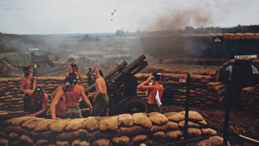 An artillery unit at work, shirtless and several clasping their ear muffs against the sound of shells being fired off.