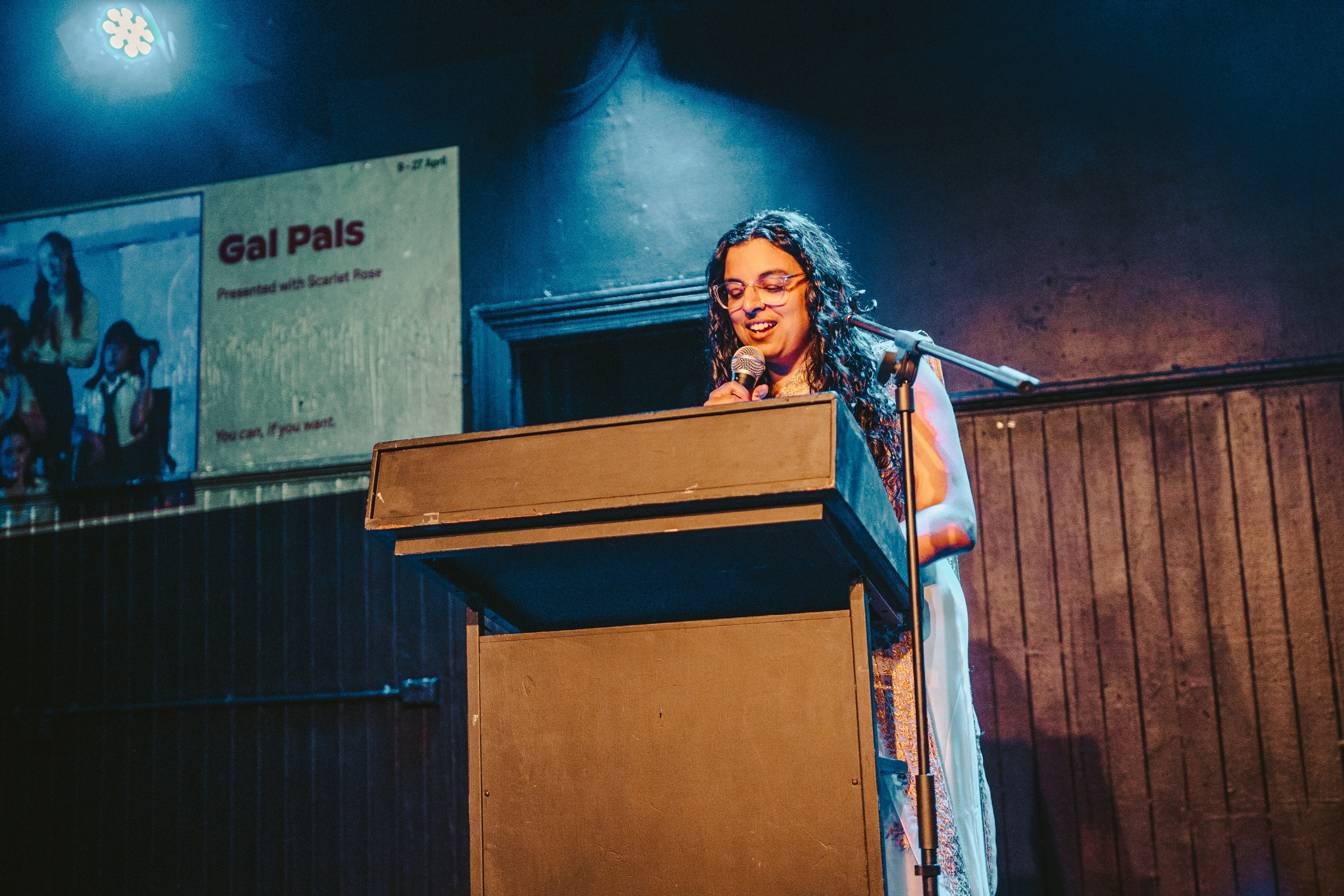Uma mulher australiana do sul da Ásia, na casa dos 20 anos, usando óculos, lê em um púlpito em uma sala azul.