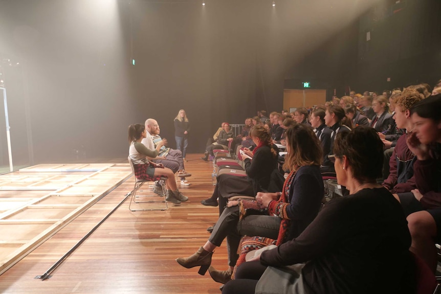 A group of students in a school theatre speak with three actors on stage