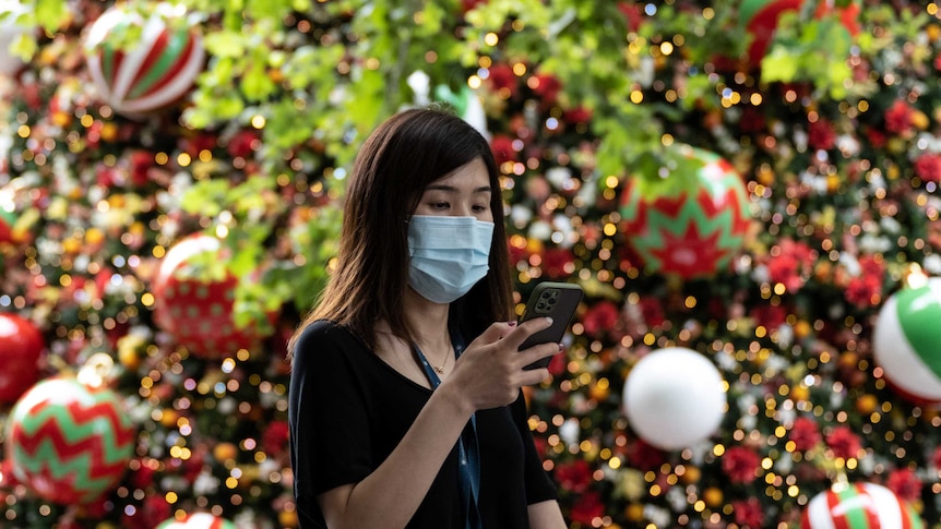 A woman in a face mask texts as she walks past a Christmas treet
