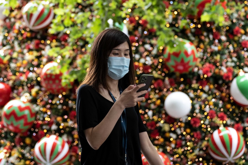 A woman in a face mask texts as she walks past a Christmas tree