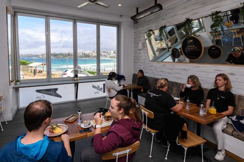 Groups of people sit at spaced out tables in a beachside cafe with views of rolling waves