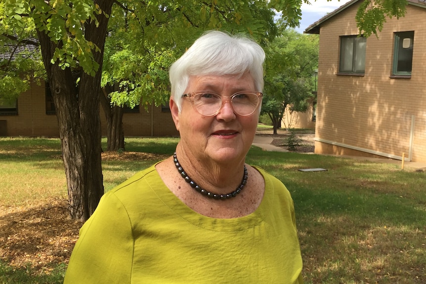 A woman with grey hair and wearing glasses and a  yellow shirt standing in front of trees