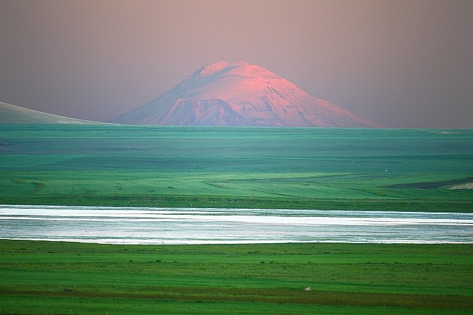 A mountian glows pink behind a vast green field and still waters.